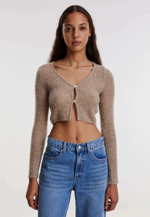 Beige kurz geschnittene Strickjacke mit langen Ärmeln, gestrickter Textur und einer Knopfleiste an der Vorderseite, kombiniert mit hochtaillierten blauen Jeans.