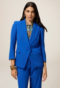 Bright blue tailored suit jacket with two buttons and notched lapels, paired with a patterned blouse. Smooth fabric with a structured fit.
