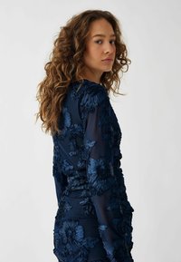 Rochie bleu marin cu broderie florală 3D, mâneci lungi și o siluetă ajustată. Prezintă un material texturat cu detalii plisate.