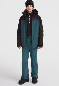 Conjunto de esquí para hombres que incluye una chaqueta impermeable negra y verde azulado, una capa intermedia negra, pantalones verdes azules y botas negras, con gafas de esquí sobre un gorro negro.