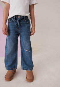 Niebieskie dżinsowe spodnie typu wide-leg z przetarciami i zniszczonym wykończeniem, noszone z brązowymi zamszowymi trzewikami na platformie i białym t-shirtem.