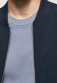 Námorní bázový bomber s plným zipem a žebrovaným límcem přes světle modrý, texturovaný svetr s kulatým výstřihem. Pohled zblízka zaměřený na oblast krku.