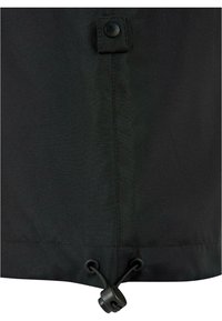 Gros plan sur l'ourlet en tissu noir avec cordon de serrage ajustable et bloqueur, plus un onglet cousu avec un bouton-pression noir au-dessus.