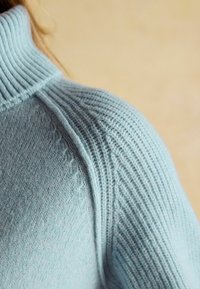 Prim-plan al unui pulover tricotat de culoare albastru deschis, cu umăr și guler înalt ribatat, pe un fundal bej.