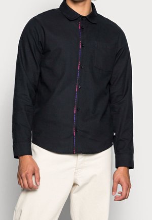 Uomo che indossa una camicia nera a maniche lunghe con bottoni, con sottili cuciture rosa e blu lungo la linea dei bottoni, e pantaloni bianchi.