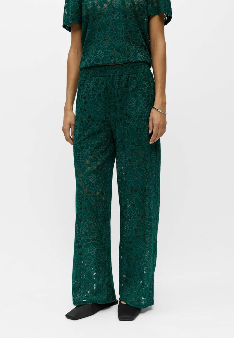 Femme portant un pantalon en dentelle florale vert foncé et un top à manches courtes assorti, associé à des chaussures noires à bout carré et un bracelet argenté.