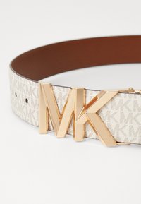 Josta ir ar krēmkrāsas auduma siksnu, uz kuras ir atkārtots logotipa raksts, un brūnu ādas aizmuguri, rotāta ar lielu zelta tonēta "MK" sprādzi.