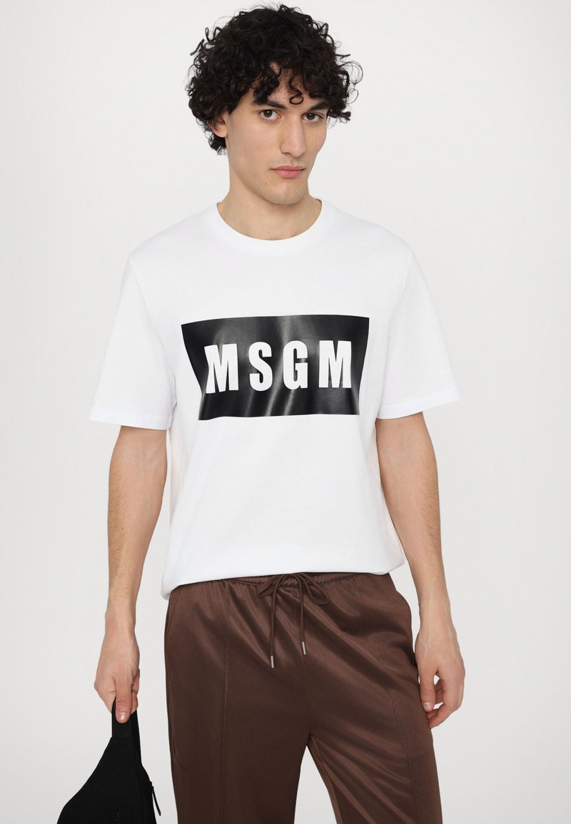 Hvid bomulds t-shirt med et stort sort rektangulært grafik med "MSGM" tekst. Matchet med brune trækknapbukser og holder en sort taske.