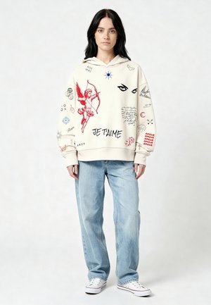 Jonge vrouw draagt een oversized crèmekleurige hoodie met rode en zwarte tekeningen, lichtblauwe jeans en witte sneakers, staand tegen een effen achtergrond.