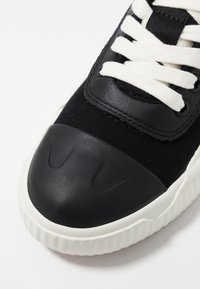 Baskets en toile noires avec un embout en caoutchouc, des lacets blancs contrastants et une semelle blanche striée. Présente une texture angulaire sur l'embout.