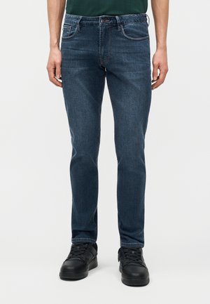 Mees, kes kannab tumesiniseid kitsaid teksaseid ja musti tenniseid, seisab ühevärvilise heleda tausta ees.