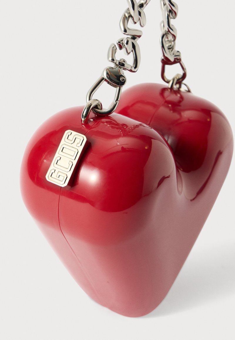 Rød hjerteformet blank taske med en sølvfarvet kædehåndtag og et metal logo-tag, der har en glat overflade og buede kanter.