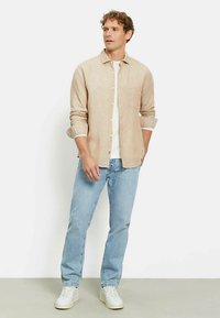 Camicia di lino beige chiaro con bottoni, abbinata a jeans di denim azzurro. Il modello indossa sneakers bianche per un look casual.