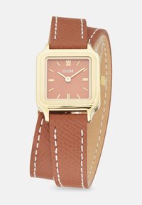 Montre carrée avec boîtier doré et cadran rose. Bracelet en cuir texturé marron avec coutures blanches. Design minimaliste avec des chiffres romains.