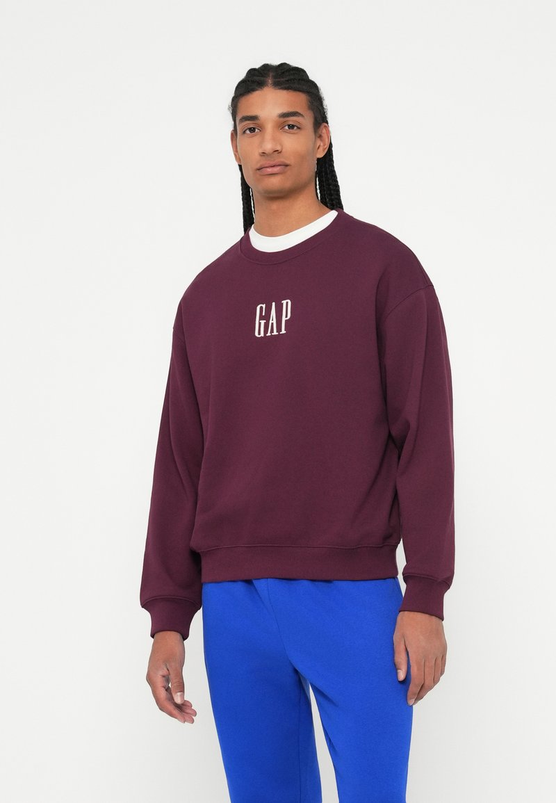Bordový sveter s bielym logom "GAP", okrúhly výstrih, rebrované manžety a lem. Kombinované s modrými teplákmi, nosené stojacim modelom.