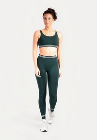 Donkergroene sportbeha en legging met hoge taille met witte gestreepte accenten. Model draagt witte sneakers en staat met de handen naast het lichaam.