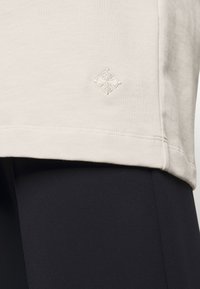Maglietta di cotone beige chiaro con un logo ricamato discreto sull'orlo. Abbinata a leggings neri che presentano una texture liscia.
