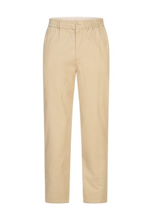 Mand iført beige bukser, hvide sneakers og en hvid skjorte, står med fødderne en smule adskilt på en ensfarvet hvid baggrund.