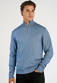 Blauwe ritstrui in een gestructureerde breisel, met lange mouwen en een hoge kraag. Het combineren met zwarte broek maakt de look compleet.