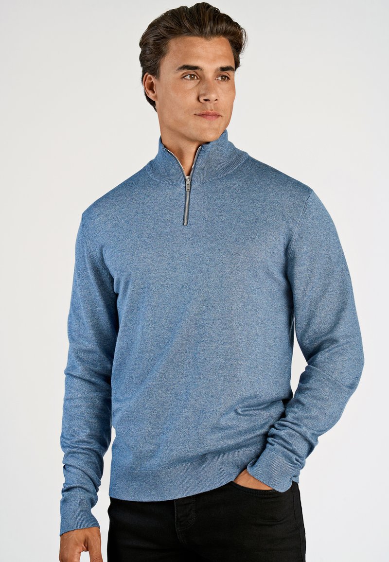 Blauwe ritstrui in een gestructureerde breisel, met lange mouwen en een hoge kraag. Het combineren met zwarte broek maakt de look compleet.