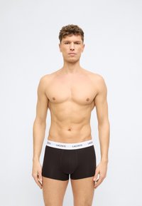 Άνδρας με ανοιχτόχρωμο δέρμα και σγουρά μαλλιά, χωρίς πουκάμισο, φοράει μαύρα εσώρουχα Lacoste με λευκή μέση, πάνω σε απλό φόντο.