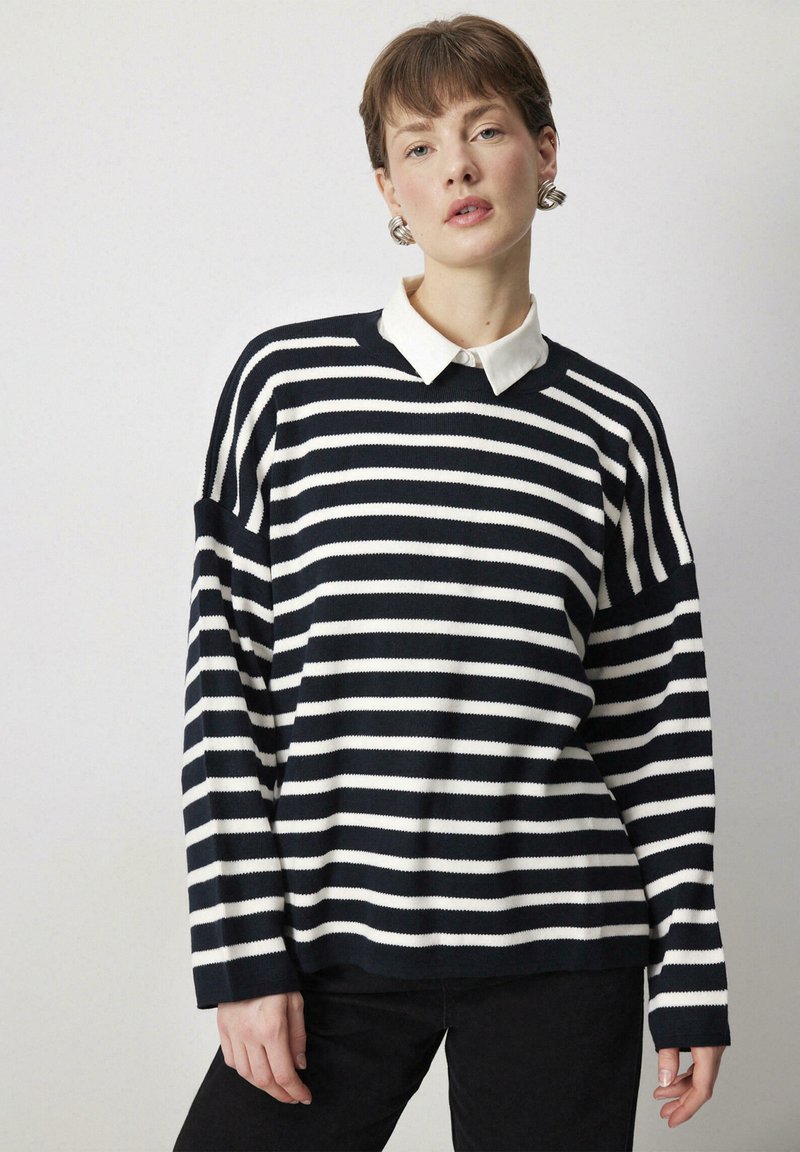 Tamsiai mėlynos ir baltos spalvos raštuotas megztinis ilgomis rankovėmis, apvaliu iškirpimu ir baltu marškiniu po juo. Minkšta megzto audinio medžiaga.