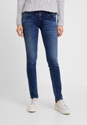 Persoon draagt donkerblauwe skinny jeans, lichtgrijze gebreide trui en witte platform sneakers, staand op een effen witte vloer.