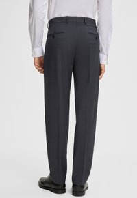 Mężczyzna w ciemnoszarych spodniach i czarnych butach, stojący tyłem do kamery na jednolitym białym tle.