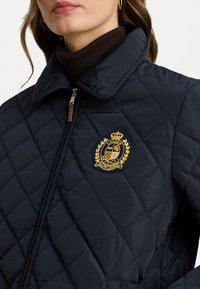 Giacca trapuntata blu navy con colletto alto, caratterizzata da un emblema dorato e una chiusura con zip. Il tessuto sembra liscio con una leggera lucentezza.