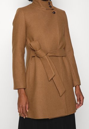 Femme portant un manteau en laine couleur camel avec un col montant, trois boutons près du cou et une ceinture nouée à la taille.