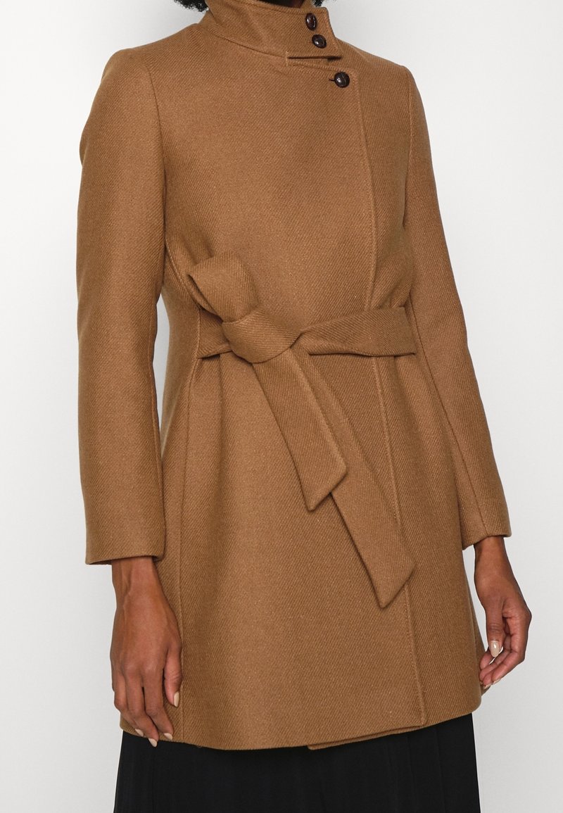 Femme portant un manteau en laine couleur camel avec un col montant, trois boutons près du cou et une ceinture nouée à la taille.