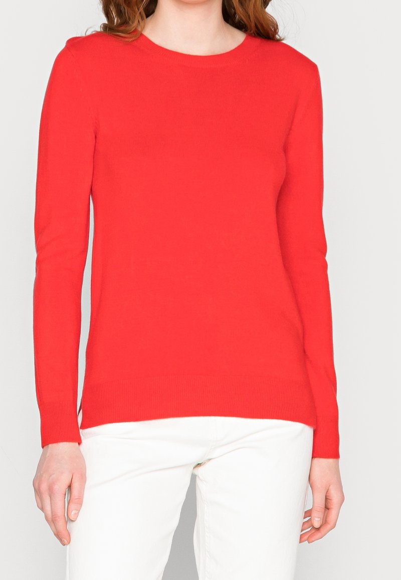 Femme portant un pull rouge à manches longues et un pantalon blanc, debout les mains relâchées le long du corps sur un fond uni.