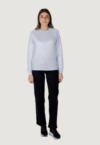 Sudadera de color azul claro con cuello redondo y puños acanalados, con un logotipo tonal en el pecho, combinada con pantalones negros de pierna recta y zapatillas negras.