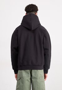 Uomo con capelli corti e ricci, che indossa un hoodie nero e pantaloni cargo verdi, rivolto di spalle su uno sfondo bianco uniforme.