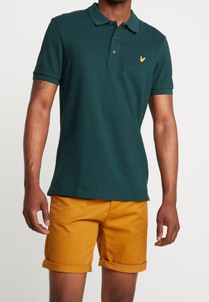 Mand iført mørkegrøn kortærmet polo skjorte med gul fuglelogo og sennepsgule optrukne shorts mod ensfarvet baggrund.