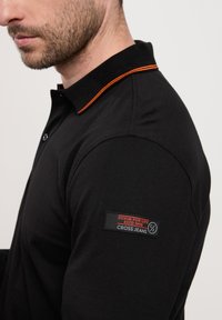 Czarna koszulka polo z kontrastowym pomarańczowym kołnierzykiem, krótkimi rękawami oraz naszywką z logo i tekstem na lewym ramieniu.