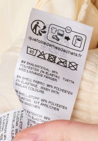 Zorglabel op een lichte stof, witte achtergrond met gedrukt tekst in meerdere talen. Bevat symbolen en wasinstructies voor materialen.