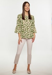 Maglietta verde con motivi a foglia crema, maniche svasate; abbinata a pantaloni attillati beige chiaro e scarpe chiare con un accento decorativo.