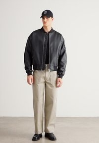 Fekete műbőr bomberdzseki, bézs bőszárú nadrág, fekete sportcipő és fekete sapka. A dzsekinek cipzárja és oldalzsebei vannak.