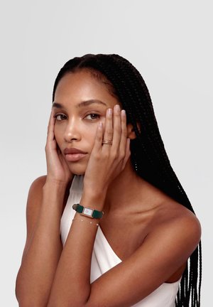 Žena s dugom pletenicom odmara lice u rukama, nosi bijelu majicu, zlatni prsten, nježnu narukvicu i pravokutni sat s zelenim kožnim remenom.