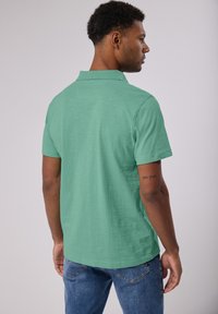 Camisa polo de manga corta en verde claro, con un cuello clásico, textura suave y un corte relajado, combinada con unos vaqueros de mezclilla.