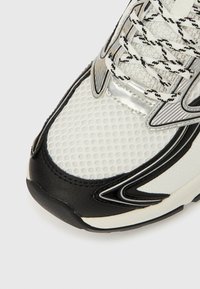 Zapatilla deportiva blanca y negra con parte superior de malla, superficie texturizada, detalles plateados y cordones con estampado. Punta redondeada y suela acolchada.