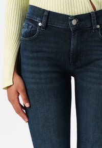Mørkeblå denim skinny jeans med midjehøyde, frontlommer og metallknappdetaljer. Stoffet har en glatt tekstur og lett stretch.