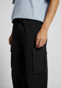 Svarta cargobyxor med flera stora sidofickor, lös passform och dragsko i midjan. Tyget verkar slätt med en matt yta.