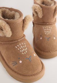 Bottes marron en faux daim avec une doublure duveteuse, un visage d'animal brodé et des oreilles texturées. Les semelles sont plates et rainurées pour une meilleure adhérence.