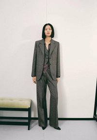 Traje gris de tejido tosco con un blazer estructurado y pantalones de pierna ancha, superpuesto a una camisa oscura con cuello. Textura suave con detalles de botones.
