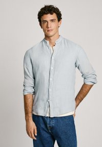 Svijetloplava, lanena košulja dugih rukava s ovratnikom bez kragne, prednjim gumbima i podvrnutim rukavima. Nošena preko bijele majice i uparena s plavim trapericama.