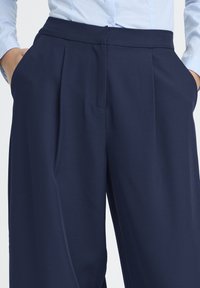 Tumesinised volditud püksid, millel on sile tekstuur ja laiad säärised. Neil on küljetaskud ja lame vöökoht, mõeldud kohandatud istuvuseks.