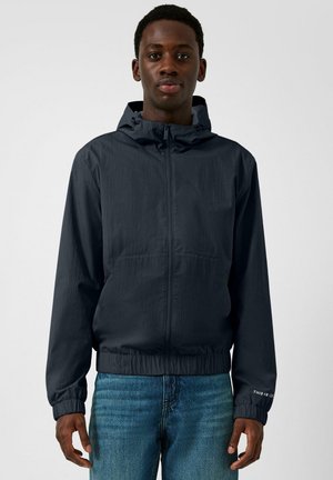 Giovane uomo visto frontalmente che indossa una giacca blu con cappuccio e zip e jeans blu su uno sfondo chiaro uniforme.