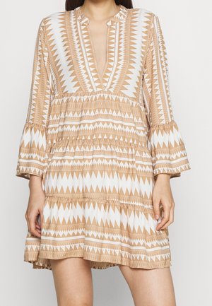 Femme portant une robe beige et blanche à motifs, manches longues, col en V et jupe à volants, debout devant un fond uni.